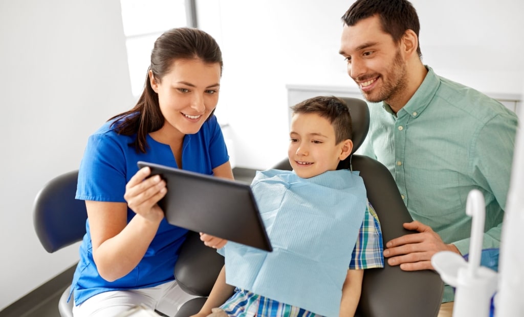 Dentist showing treatment details on a tablet to a child and parent, illustrating Dental Revenue Cycle Management in action.